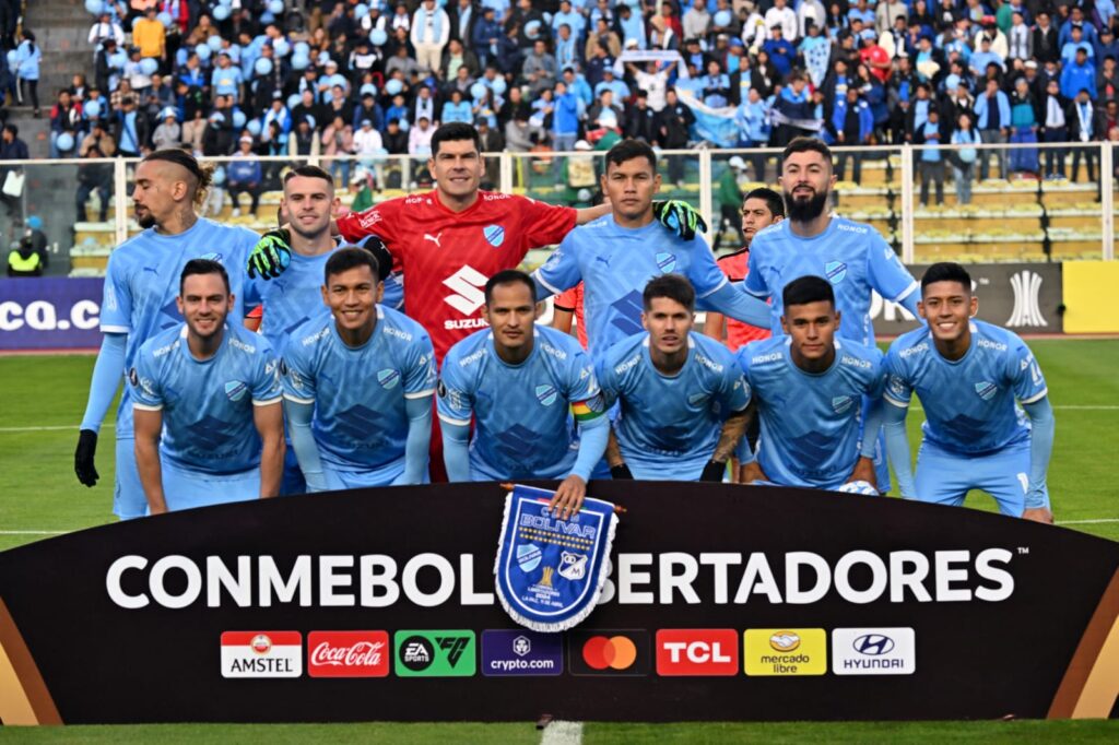 Time do Bolívar, adversário do Flamengo na Libertadores, com brasão de jogo e perfilado antes de partida pelo torneio continental com torcedores ao fundo e pedaço da arquibancada de seu estádio aparecendo