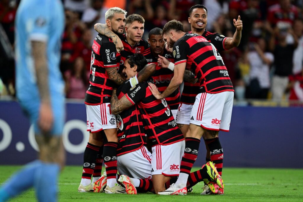 Jogadores do Flamengo comemoram gol contra o Bolívar, no Maracanã, pela Libertadores, reunidos e se abraçando no campo ao fundo da foto; atleta do Bolívar caminha desolado na frente dos rubro-negros